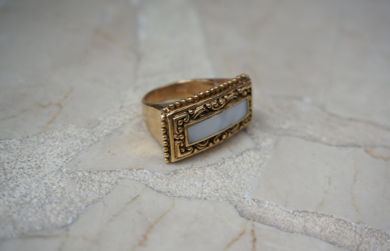 May include: A gold ring with a rectangular white stone set in a detailed ornate frame.