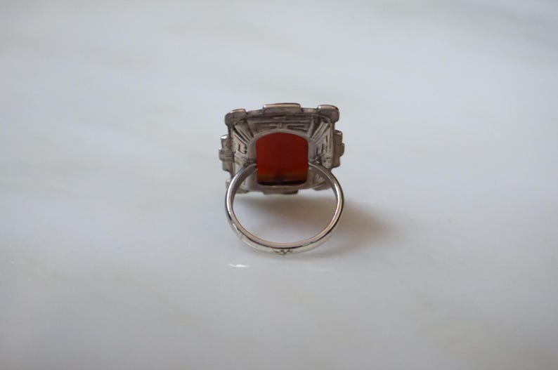 May include: A silver ring with a square-shaped red gemstone set in a geometric design.