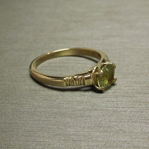 May include: A gold ring with a green gemstone set in a prong setting. The band is textured with a series of small ridges.