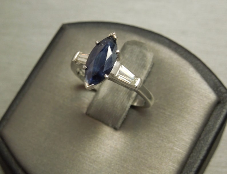 以下が含まれることがあります： 白金にマーキスカットの青いサファイアがセットされた婚約指輪。バンドには小さなダイヤモンドが2つ付いています。
