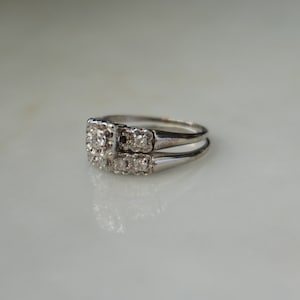May include: A silver engagement ring set with a square center stone and smaller surrounding stones. The matching band sits below the engagement ring. The rings are on a white surface.
