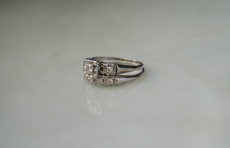 May include: A silver engagement ring set with a square center stone and smaller surrounding stones. The matching band sits below the engagement ring. The rings are on a white surface.