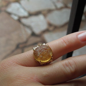 Op de afbeelding: Een gouden ring met een grote, ronde, gele edelsteen. De edelsteen is gegraveerd met een gedetailleerd ontwerp.