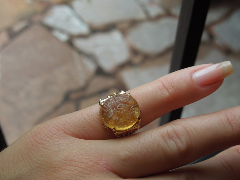 Op de afbeelding: Een gouden ring met een grote, ronde, gele edelsteen. De edelsteen is gegraveerd met een gedetailleerd ontwerp.