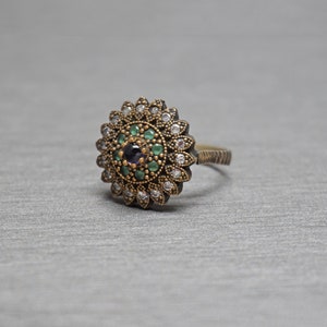 Pu&ograve; includere: Un anello d'argento con una grande montatura rotonda multi-pietra. La montatura presenta una pietra centrale viola circondata da pietre verdi e bianche pi&ugrave; piccole.