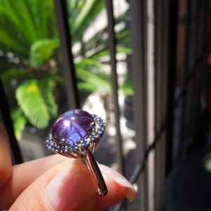 May include: A silver ring featuring a large, round, purple gemstone with a star-like effect. The gemstone is surrounded by smaller, blue gemstones. The ring is held up in front of a window with green foliage visible in the background.