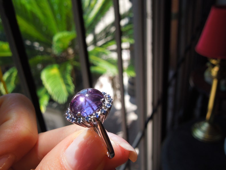 May include: A silver ring featuring a large, round, purple gemstone with a star-like effect. The gemstone is surrounded by smaller, blue gemstones. The ring is held up in front of a window with green foliage visible in the background.