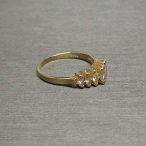 May include: A gold ring featuring a row of five oval-cut gemstones. The ring has a polished gold band and the gemstones are set close together. The ring is displayed on a gray surface.