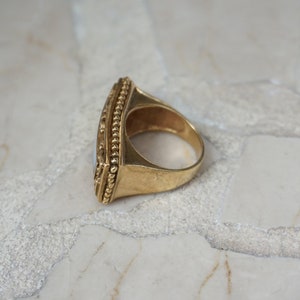 May include: A gold ring with a large, rectangular, opaque stone set in a bezel setting. The ring has a beaded edge and a wide band.