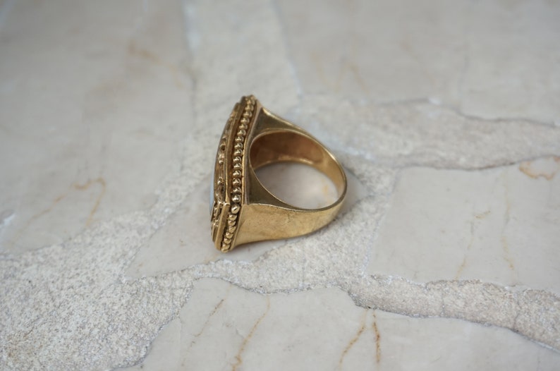 May include: A gold ring with a large, rectangular, opaque stone set in a bezel setting. The ring has a beaded edge and a wide band.