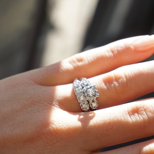 May include: A close-up of a hand wearing a diamond engagement ring and a matching wedding band. The rings are set with multiple small, clear diamonds. The hand is in soft focus, with sunlight illuminating the jewelry.
