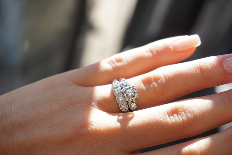 May include: A close-up of a hand wearing a diamond engagement ring and a matching wedding band. The rings are set with multiple small, clear diamonds. The hand is in soft focus, with sunlight illuminating the jewelry.