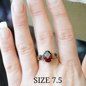 May include: A gold ring with a large red gemstone and two smaller brown gemstones. The ring is on a hand, and the text "SIZE 7.5" is visible below the ring.