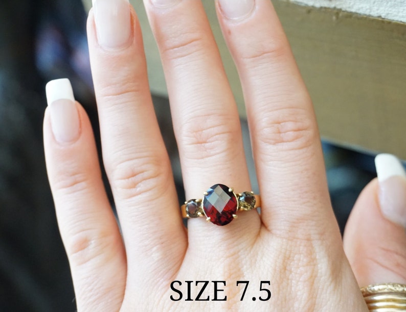 May include: A gold ring with a large red gemstone and two smaller brown gemstones. The ring is on a hand, and the text "SIZE 7.5" is visible below the ring.