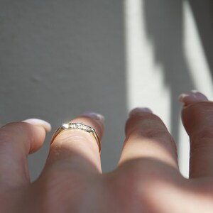 May include: A gold ring with small, clear stones set across the top. The ring is on a finger, with the hand and other fingers visible. The background is a light grey with shadows.