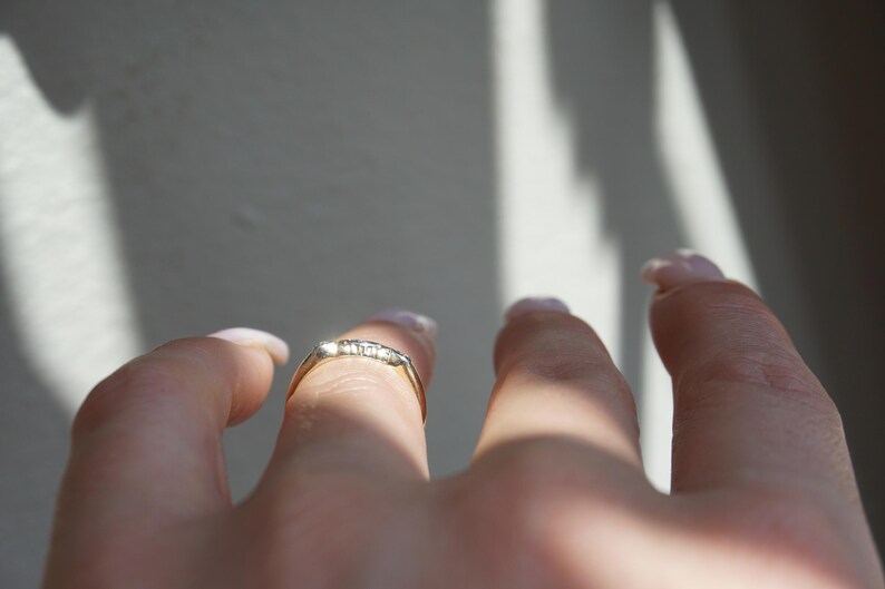 May include: A gold ring with small, clear stones set across the top. The ring is on a finger, with the hand and other fingers visible. The background is a light grey with shadows.