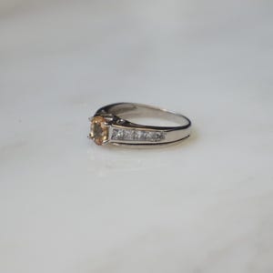 Pode incluir: Um anel de prata com uma gema oval de cor castanho claro e uma banda de pequenas gemas transparentes.