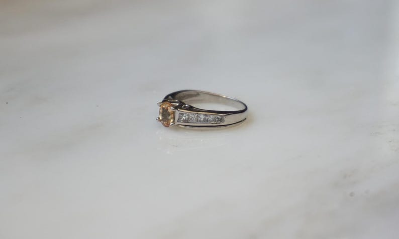 Pode incluir: Um anel de prata com uma gema oval de cor castanho claro e uma banda de pequenas gemas transparentes.