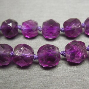 Pode incluir: Um fio de contas de ametista roxa, cada conta &eacute; facetada e tem uma forma geom&eacute;trica. As contas s&atilde;o enfiadas em um cord&atilde;o roxo.