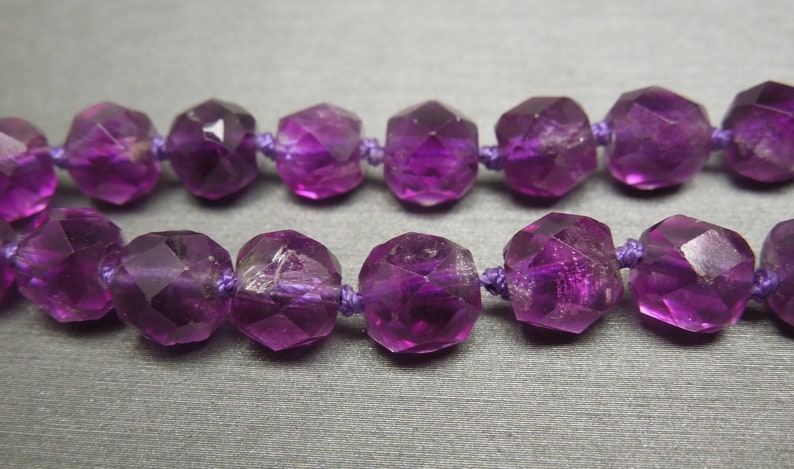 Pode incluir: Um fio de contas de ametista roxa, cada conta &eacute; facetada e tem uma forma geom&eacute;trica. As contas s&atilde;o enfiadas em um cord&atilde;o roxo.