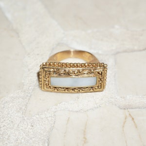 May include: A gold ring with a rectangular white mother-of-pearl inlay. The ring has a detailed ornate design around the inlay.