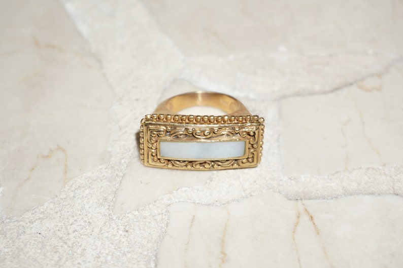 May include: A gold ring with a rectangular white mother-of-pearl inlay. The ring has a detailed ornate design around the inlay.