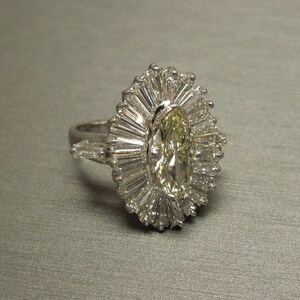 Op de afbeelding: Een zilveren ring met een grote, ovale, gele diamant omringd door kleinere diamanten in een zonnestralenpatroon.