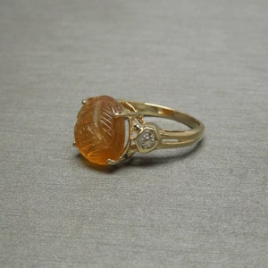 May include: A gold ring with an oval-shaped orange gemstone set in a carved design. A smaller diamond is set on the side of the ring.