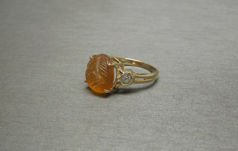 May include: A gold ring with an oval-shaped orange gemstone set in a carved design. A smaller diamond is set on the side of the ring.