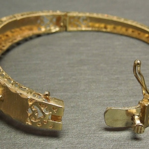 May include: A gold-toned bangle bracelet with a delicate filigree design and a hinged clasp. The bracelet is adorned with small, white opal stones.