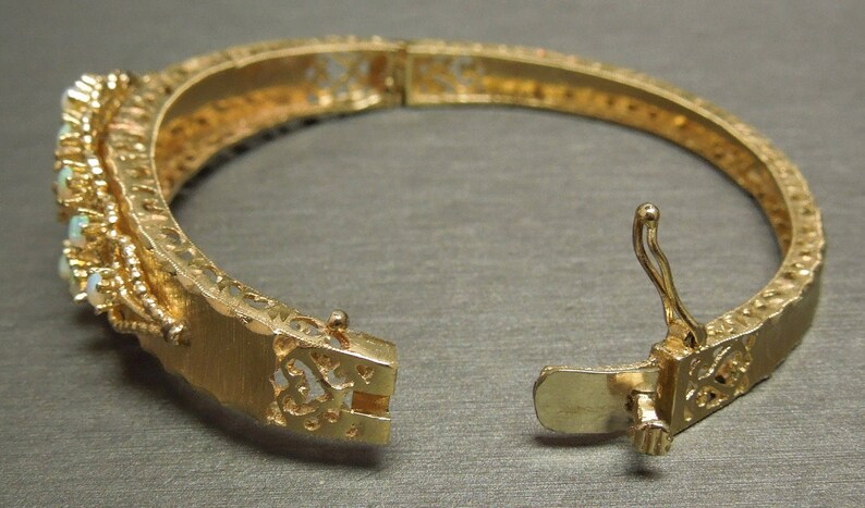 May include: A gold-toned bangle bracelet with a delicate filigree design and a hinged clasp. The bracelet is adorned with small, white opal stones.