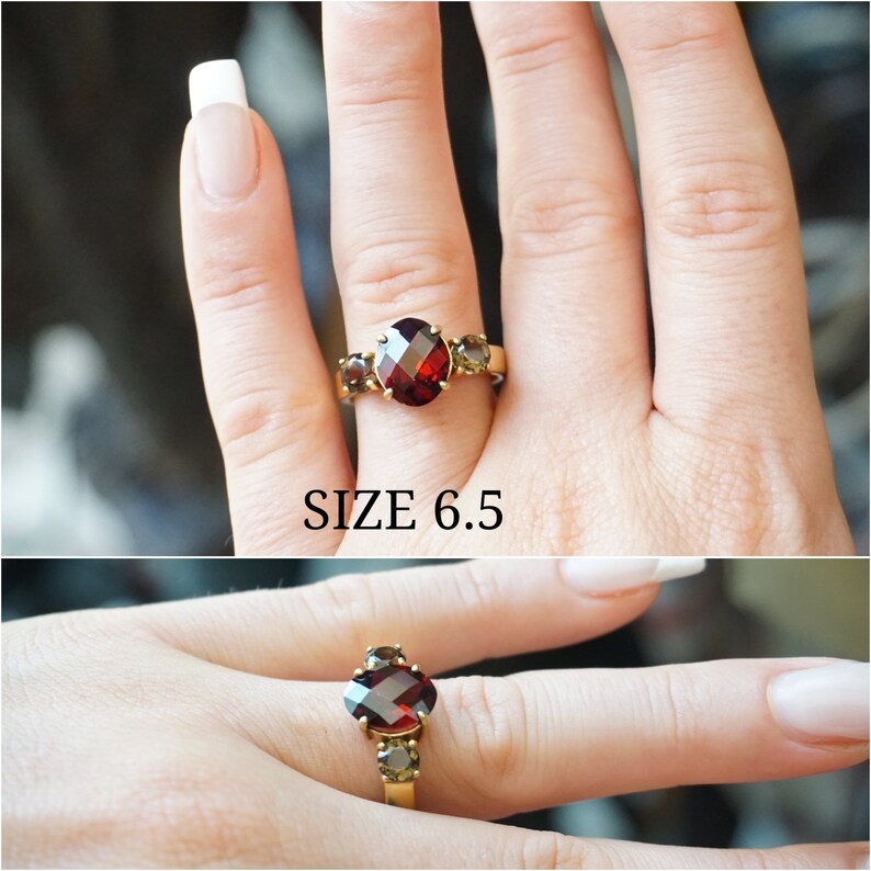 May include: A gold ring with a large red oval gemstone and two smaller brown gemstones. The ring is shown on a finger, and the text "SIZE 6.5" is visible below the ring.