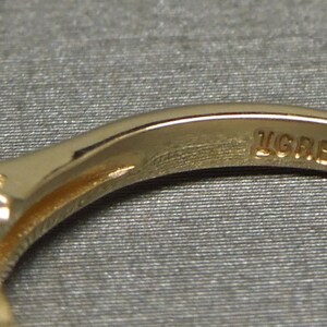 May include: Close-up of a gold ring with a row of small, sparkling stones. The ring's band is smooth and polished, with the inscription "LGRB" visible. The ring is set against a neutral, textured background.