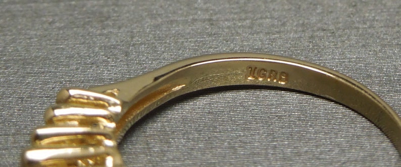May include: Close-up of a gold ring with a row of small, sparkling stones. The ring's band is smooth and polished, with the inscription "LGRB" visible. The ring is set against a neutral, textured background.