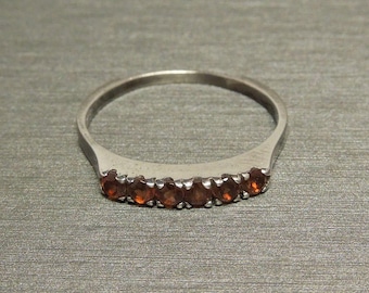 May include: A silver ring with a row of six small, round, orange-red gemstones. The ring band is smooth and the gemstones are set close together. The ring is on a textured, gray surface.
