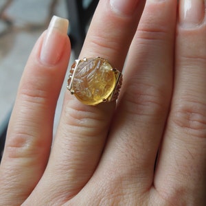 Op de afbeelding: Een gouden ring met een grote, ronde, gele edelsteen. De edelsteen heeft een gegraveerd ontwerp en is gezet in een gouden band.