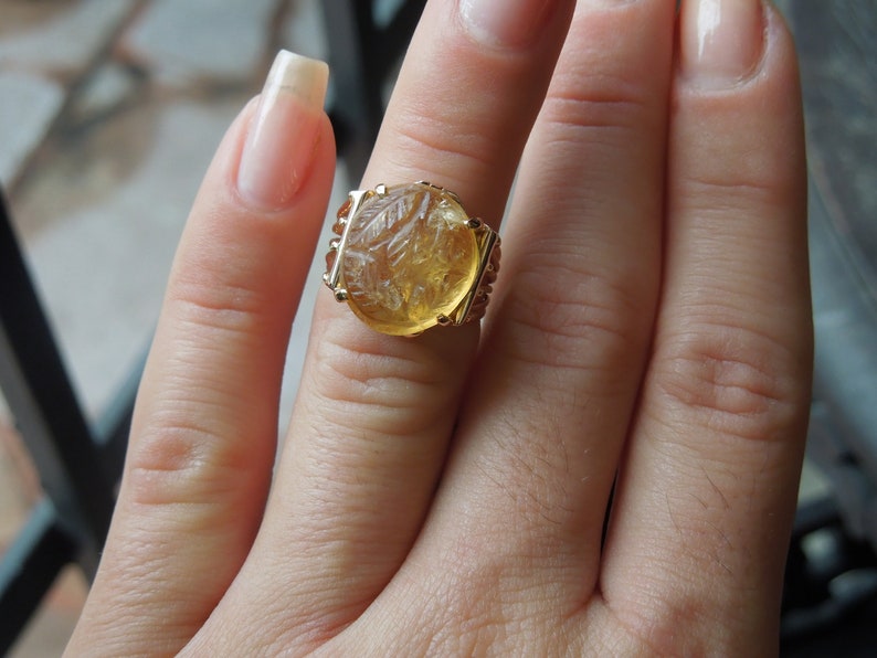 Op de afbeelding: Een gouden ring met een grote, ronde, gele edelsteen. De edelsteen heeft een gegraveerd ontwerp en is gezet in een gouden band.