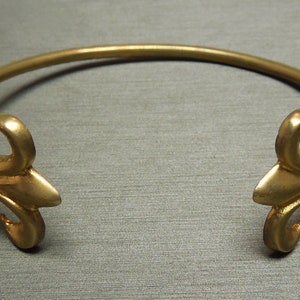 May include: A gold-toned metal bracelet with two fleur-de-lis designs on the ends.