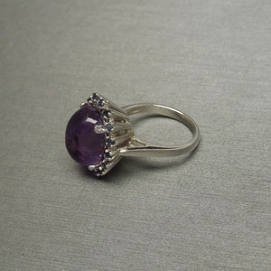 May include: A silver ring featuring a large, oval-cut, deep purple amethyst gemstone. The amethyst is surrounded by smaller, dark blue gemstones. The ring has a classic, elegant design.
