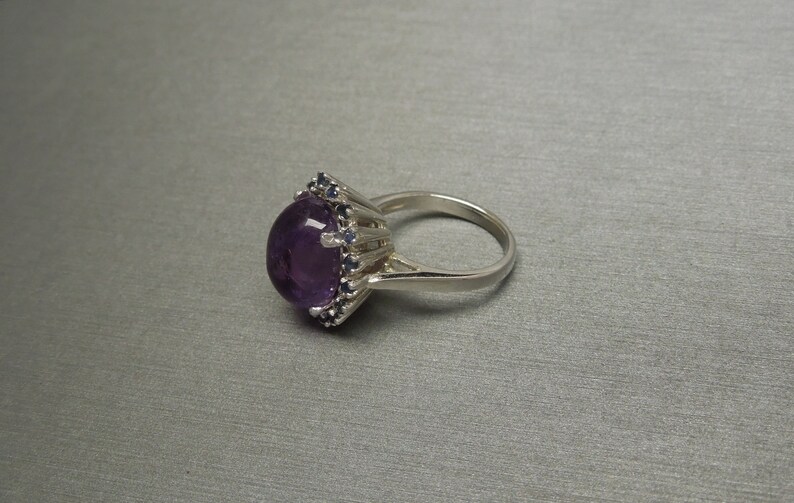 May include: A silver ring featuring a large, oval-cut, deep purple amethyst gemstone. The amethyst is surrounded by smaller, dark blue gemstones. The ring has a classic, elegant design.