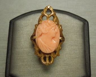 May include: A gold-toned cameo ring featuring a delicate carved portrait of a woman in a pale pink cameo. The ring has an intricate filigree design.