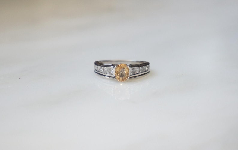 Pode incluir: Um anel de prata com uma &uacute;nica pedra preciosa de top&aacute;zio de forma oval e uma banda de diamantes menores.