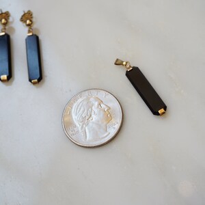 以下が含まれることがあります： 金色のアクセントが付いた黒オニキスのイヤリングのペアと、金色のアクセントが付いた黒オニキスのペンダント。ジュエリーは、サイズ比較のために米国の25セント硬貨の隣に置かれています。