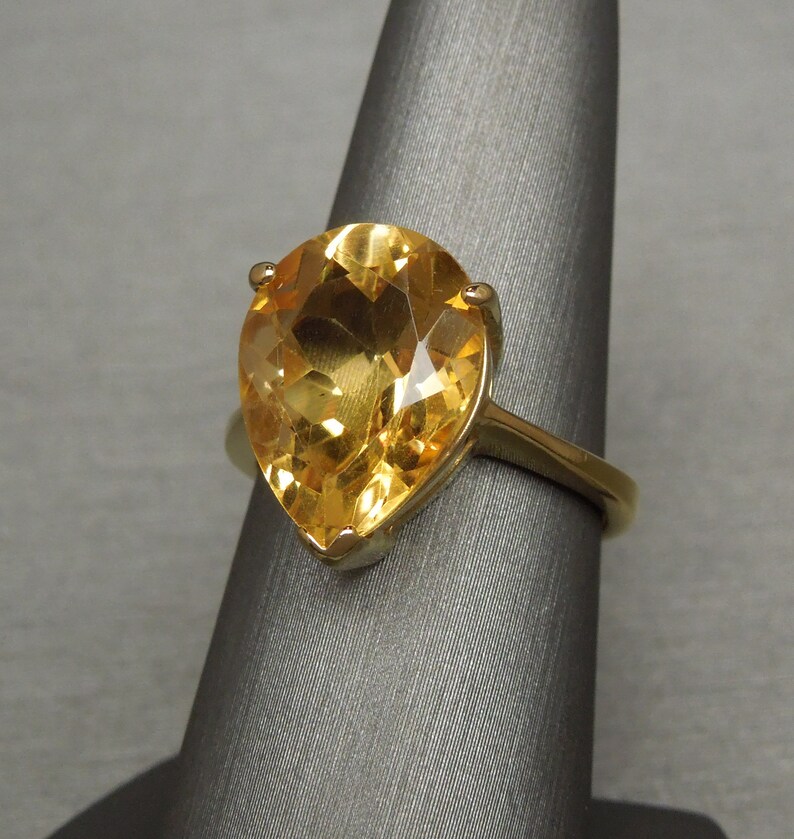 May include: A gold ring featuring a large pear-shaped topaz gemstone. The gemstone is a bright yellow colour.