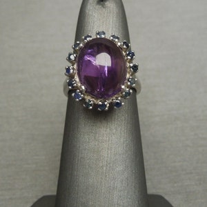May include: A silver ring featuring a large, oval-shaped, polished purple gemstone, surrounded by a halo of smaller, round, blue gemstones. The ring is displayed on a gray stand, showcasing its design and color contrast.