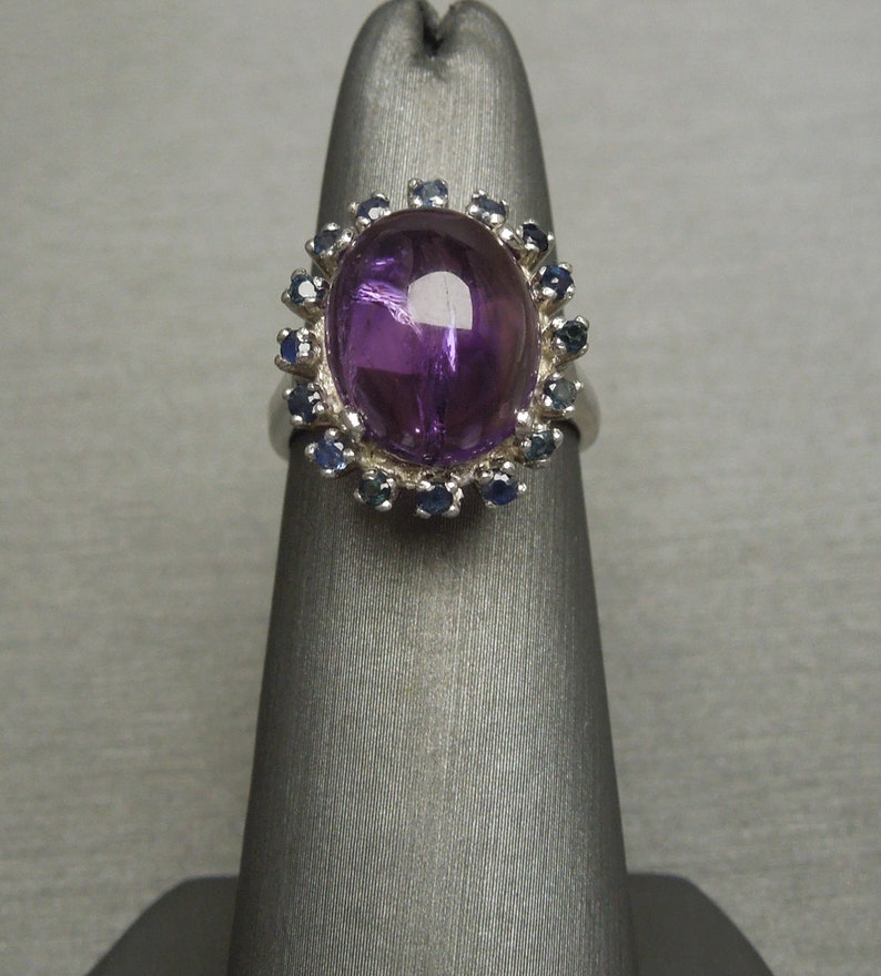 May include: A silver ring featuring a large, oval-shaped, polished purple gemstone, surrounded by a halo of smaller, round, blue gemstones. The ring is displayed on a gray stand, showcasing its design and color contrast.