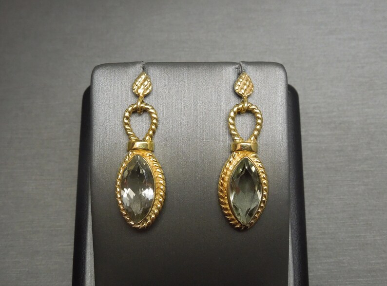 May include: A pair of gold earrings with a green gemstone in the centre of each earring. The earrings are shaped like a teardrop and have a delicate design.
