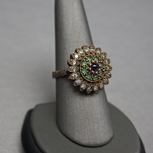 Pu&ograve; includere: Un anello d'argento con un grande designo floreale rotondo. Il centro del design presenta una pietra preziosa blu circondata da pietre preziose verdi e pietre preziose bianche pi&ugrave; piccole.