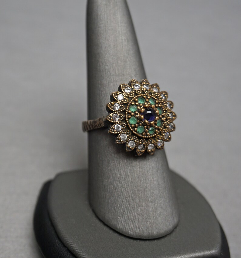 Pu&ograve; includere: Un anello d'argento con un grande designo floreale rotondo. Il centro del design presenta una pietra preziosa blu circondata da pietre preziose verdi e pietre preziose bianche pi&ugrave; piccole.