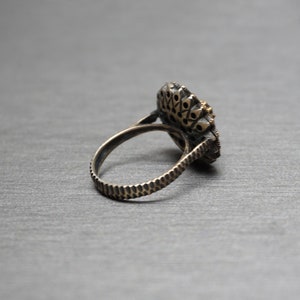 Pu&ograve; includere: Un anello d'argento con un grande designo ornato nero e oro. L'anello &egrave; posto su una superficie grigia.